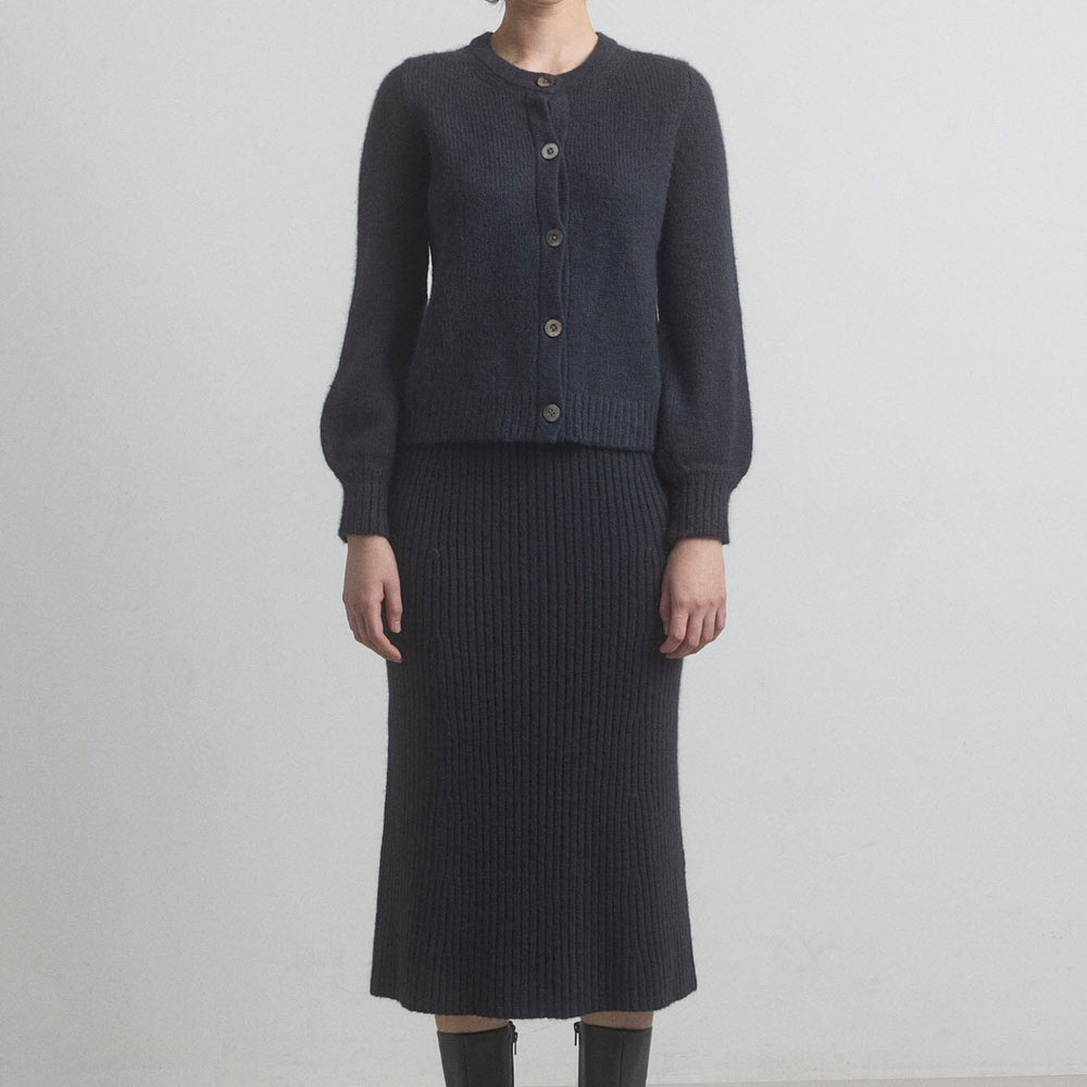 Woman wearing a dark gray cardigan and black ribbed merino wool skirt set on a plain background