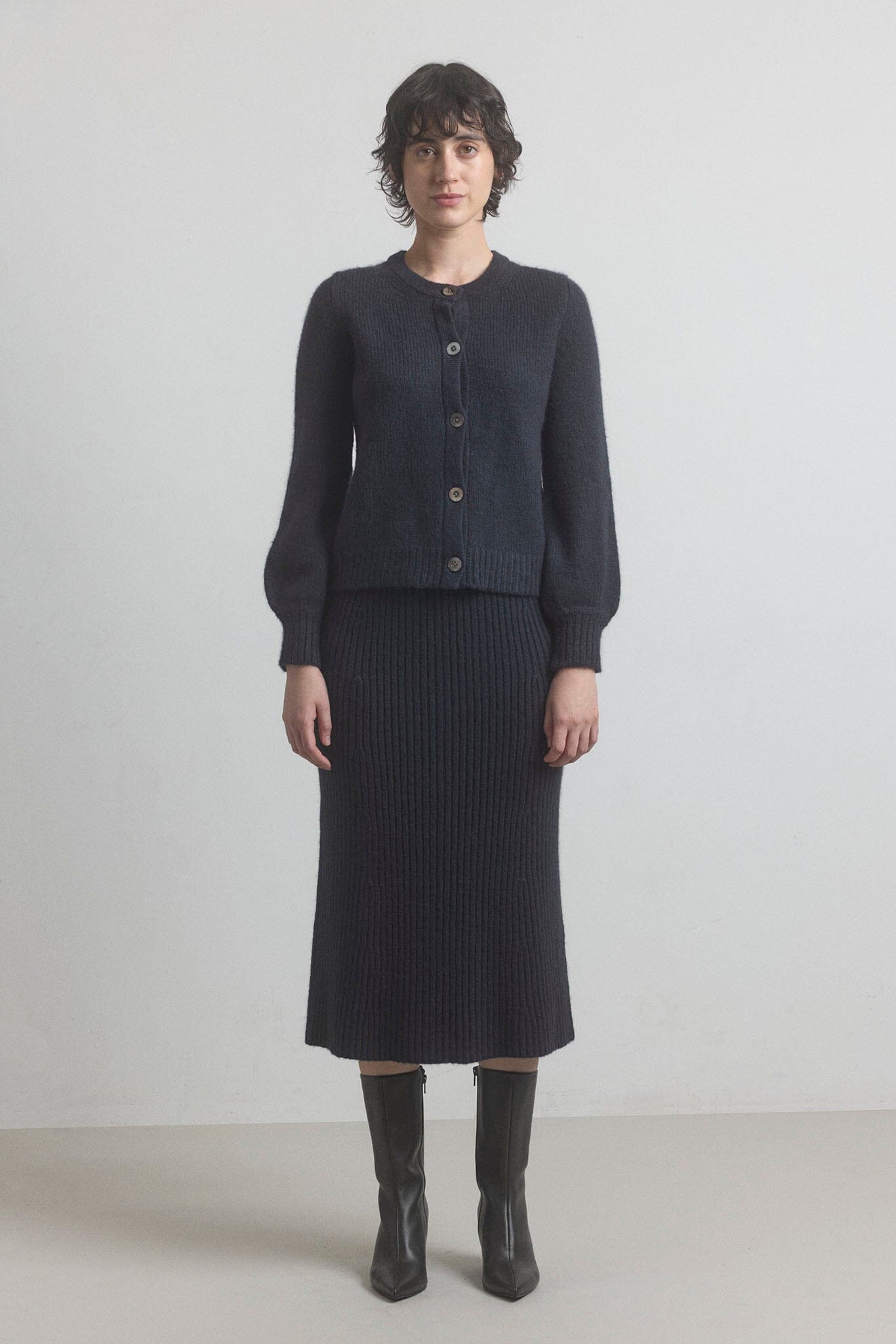 Woman wearing a dark gray cardigan and black ribbed merino wool skirt set on a plain background