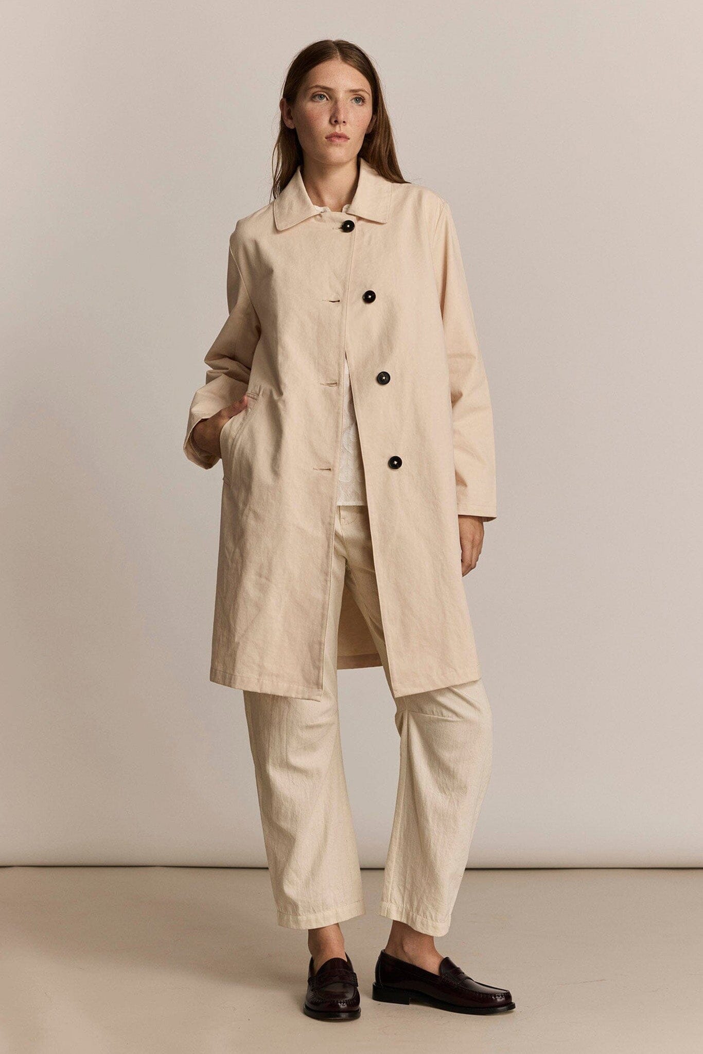 Woman wearing a beige trench coat and pants against a plain background