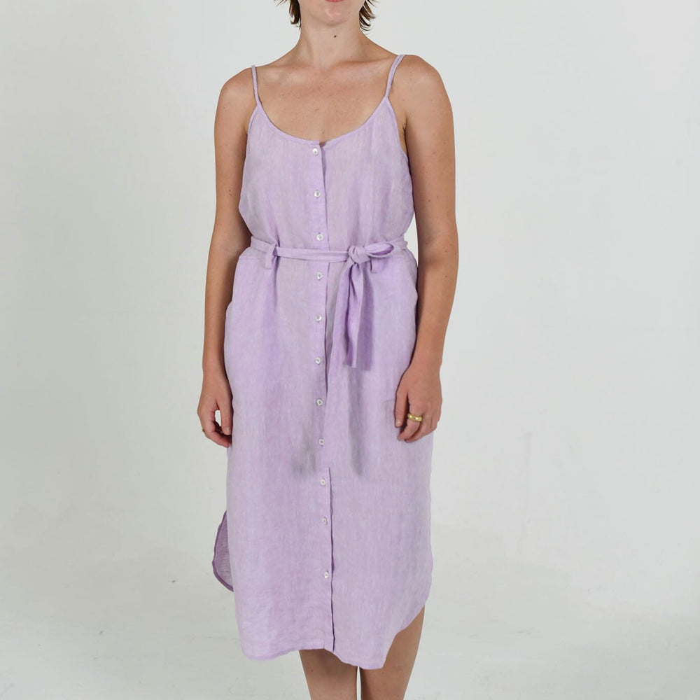 A woman standing in a studio setting, wearing a lavender sleeveless dress with a scoop neckline, half button placket, and adjustable straps. The dress has front slash pockets and a deep shirttail hem, accompanied by a belt.