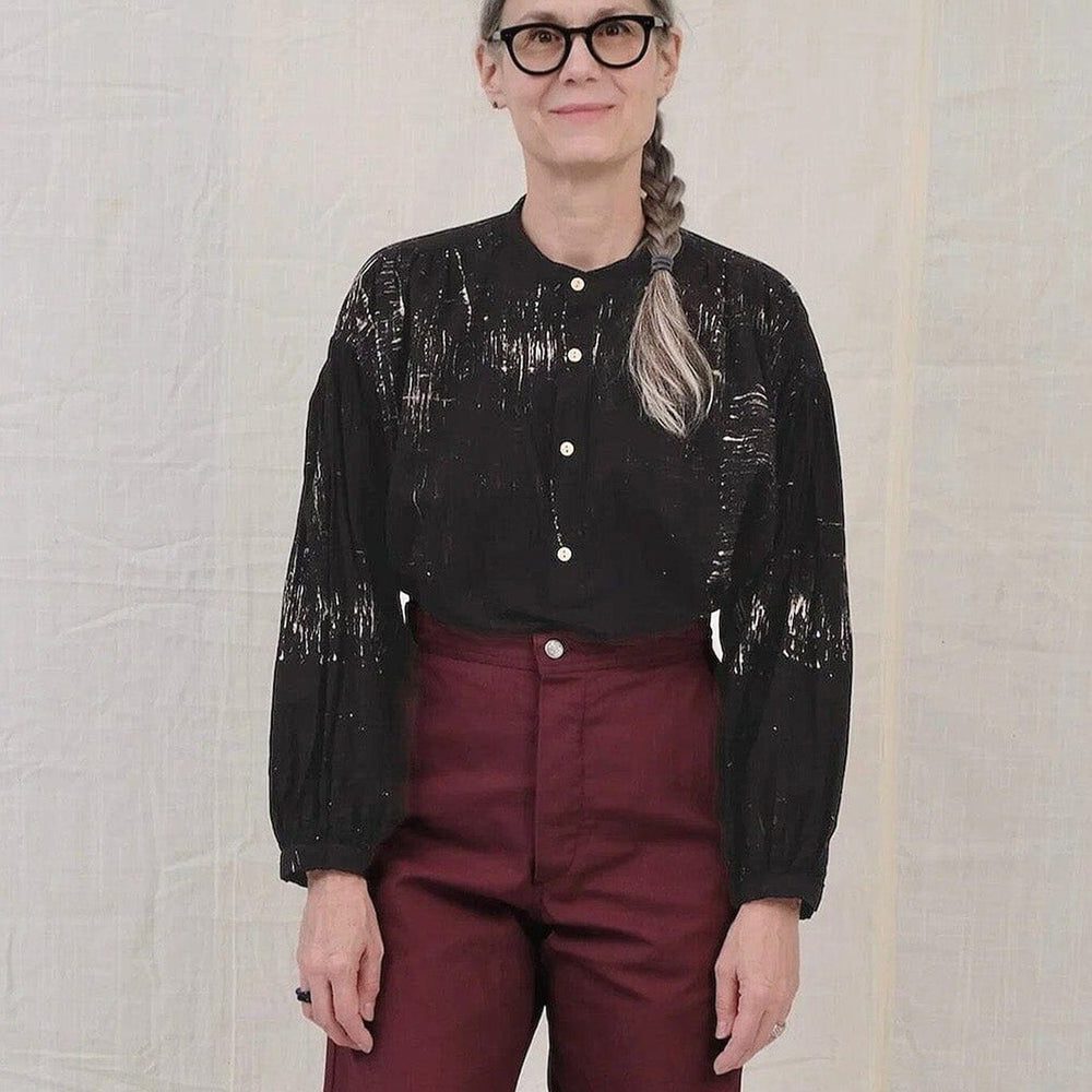 
                  
                    A woman wearing a black silk blouse with white painted pattern and burgundy pants
                  
                