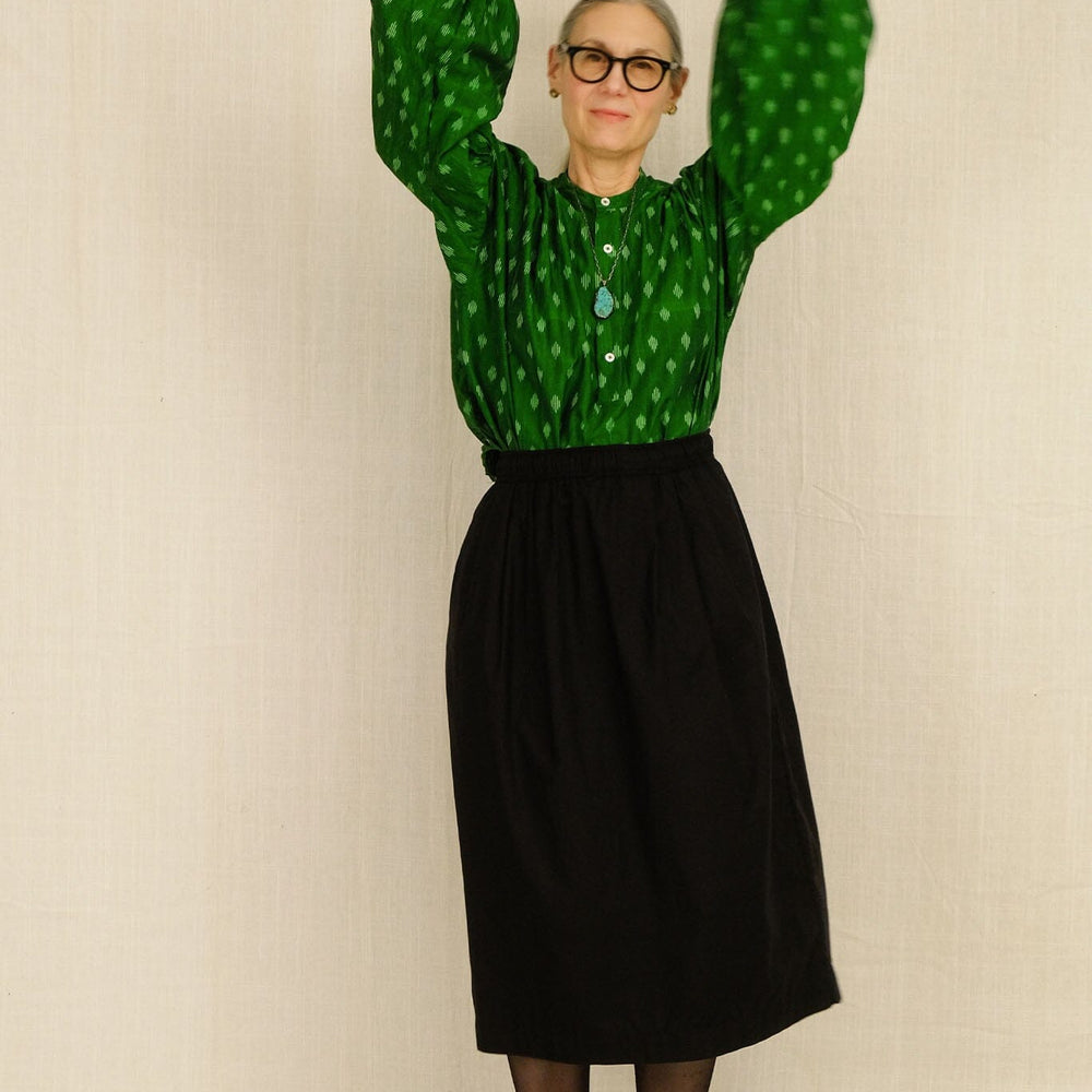 Woman wearing a green patterned long sleeve top and black midi skirt against a beige background