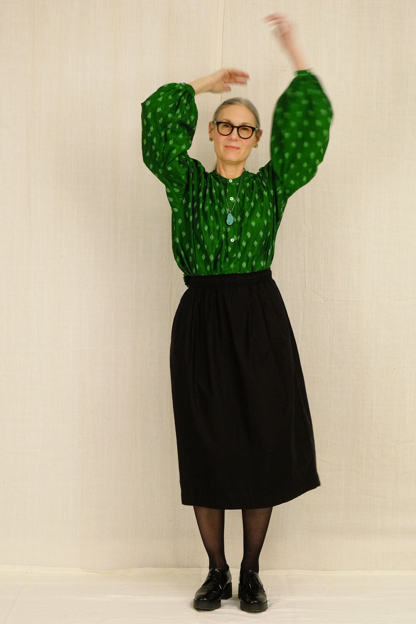 Woman wearing a green patterned long sleeve top and black midi skirt against a beige background