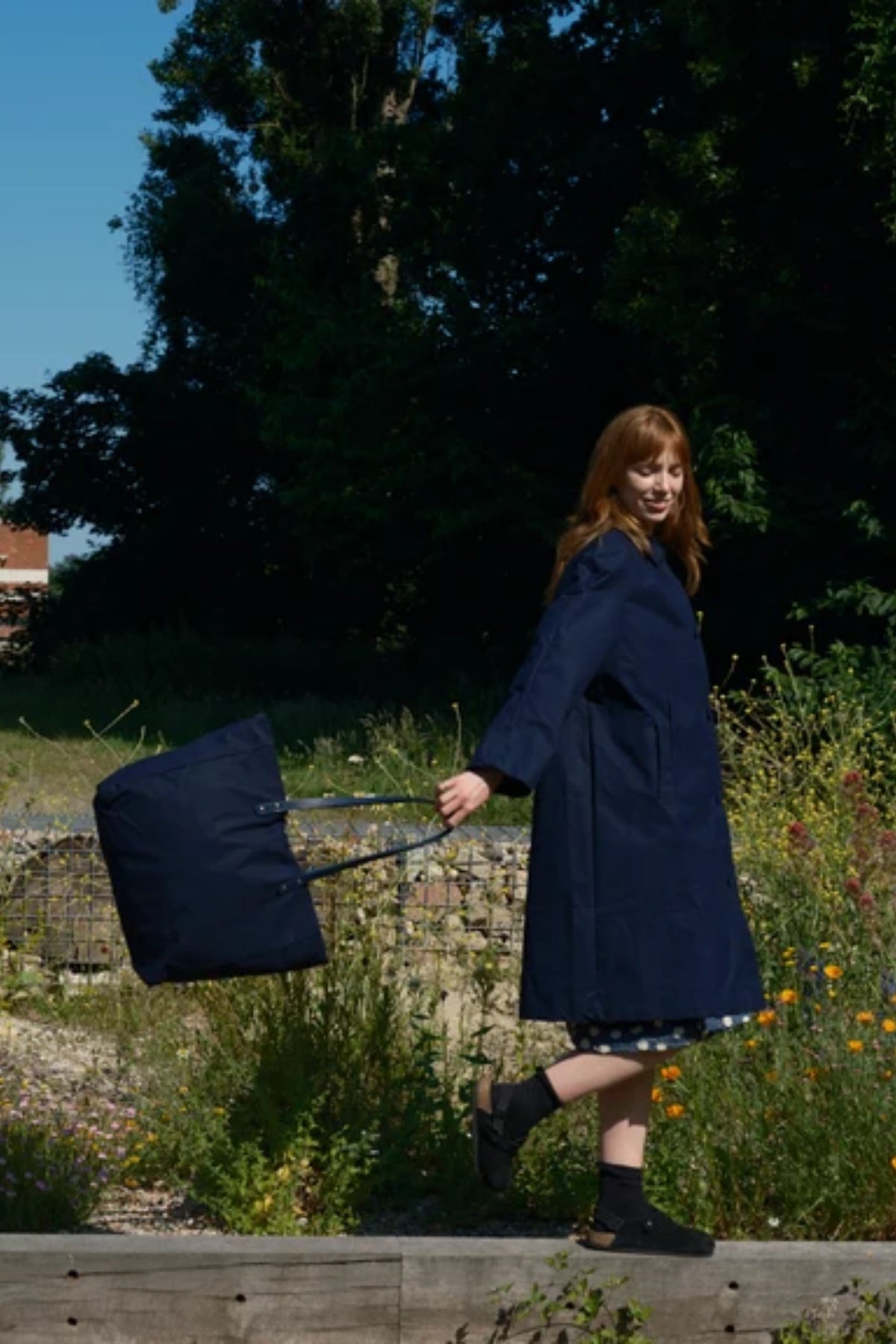 A woman wearing a long navy coat walks outdoors while pulling a matching navy duffel bag with a telescoping handle. The scene is set in a grassy, wooded outdoor area with a wooden fence in the foreground.