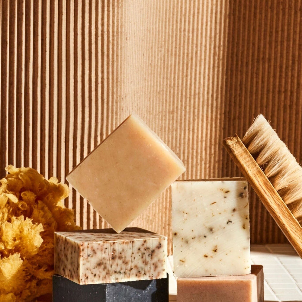 A variety of bars of soap with a beige, brown, and black color palette, displayed alongside a brush and what appears to be dried flowers.