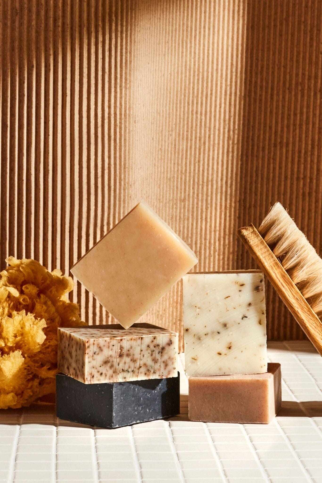 A variety of bars of soap with a beige, brown, and black color palette, displayed alongside a brush and what appears to be dried flowers.