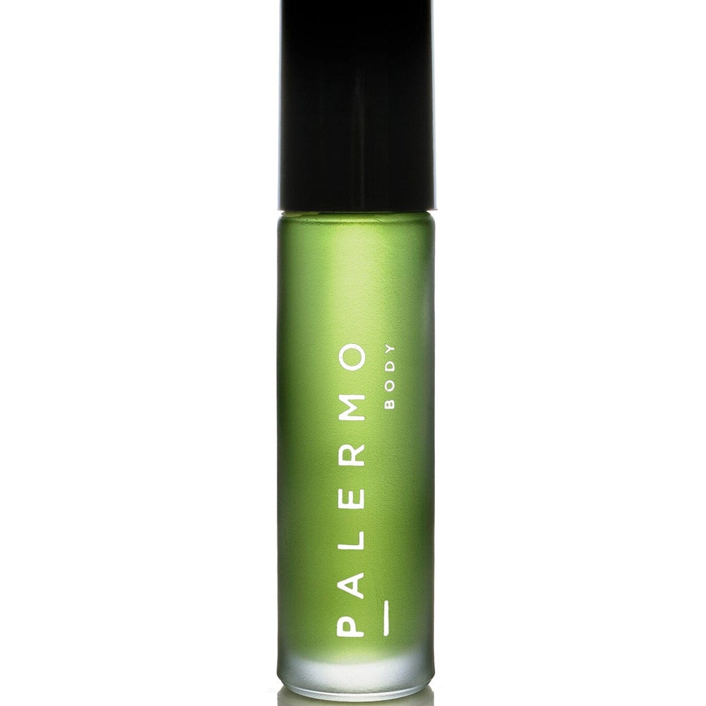 A bottle of Palermo Body Sacred Aromatherapy Oil with a green label, placed on a surface with a smoky backdrop, suggesting the oil is used for aromatherapy. There are dried plant elements nearby, indicating the natural ingredients of the oil.