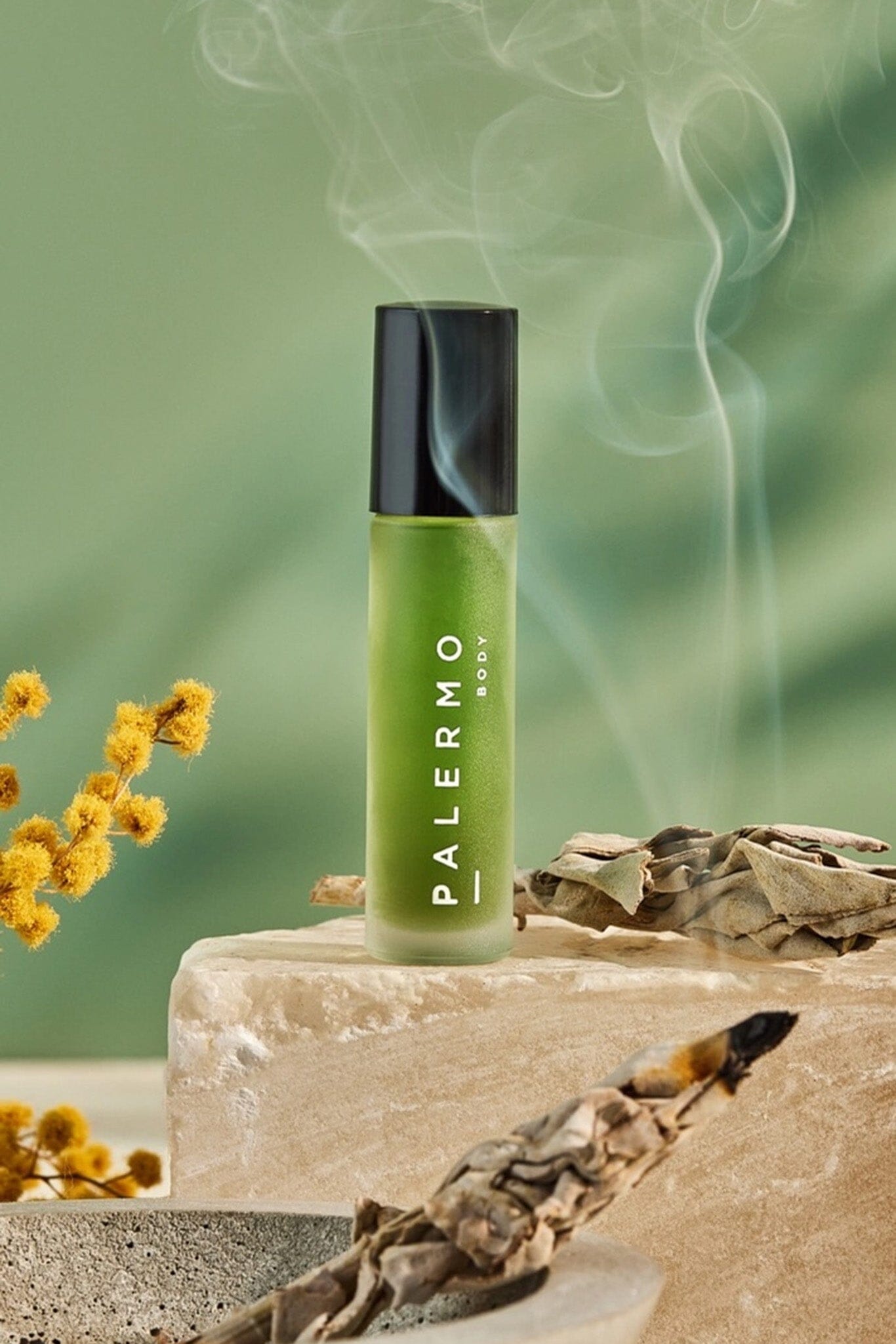 A bottle of Palermo Body Sacred Aromatherapy Oil with a green label, placed on a surface with a smoky backdrop, suggesting the oil is used for aromatherapy. There are dried plant elements nearby, indicating the natural ingredients of the oil.