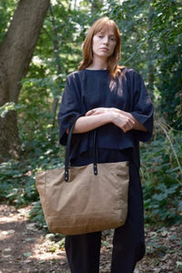 A woman standing in a wooded outdoor setting, holding a large tan canvas tote bag with black shoulder straps. She is wearing a dark navy top and dark pants.