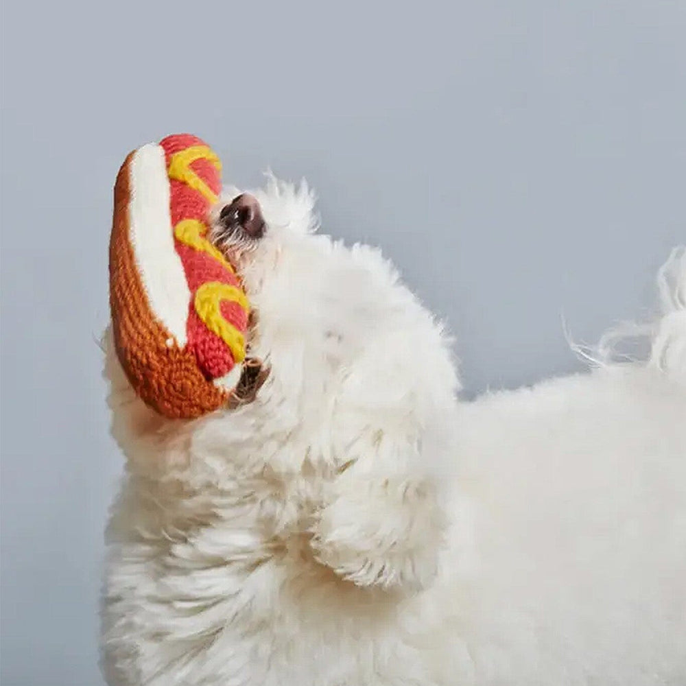 White dog playing with a hot dog-shaped toy against a light blue background