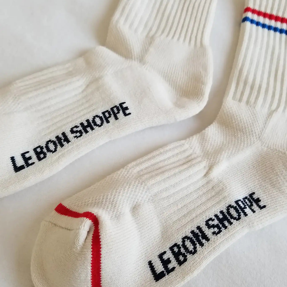 
                  
                    A close up shot of Le Bon Shoppe white ribbed cotton socks with a red and blue top stripe a logo on the bottom
                  
                