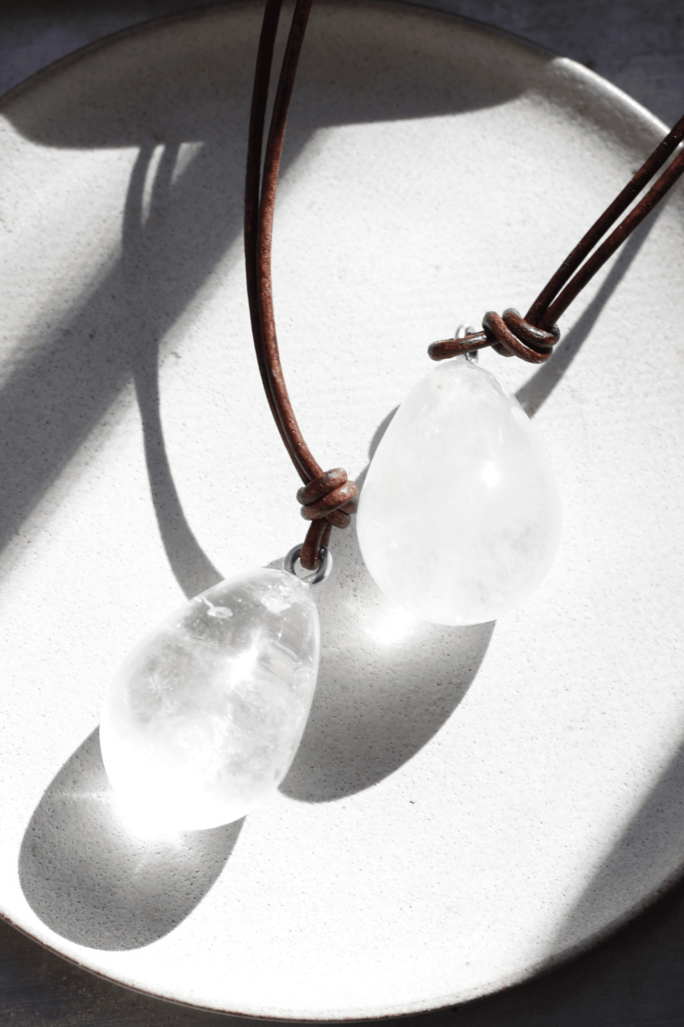 A brown leather cord necklace with three rock crystal pendants, displayed on a white plate in a shadowed setting.