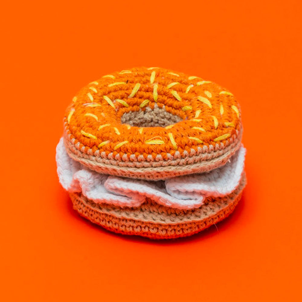 A crochet dog toy designed to resemble a bagel, with orange and white colors, on an orange background.