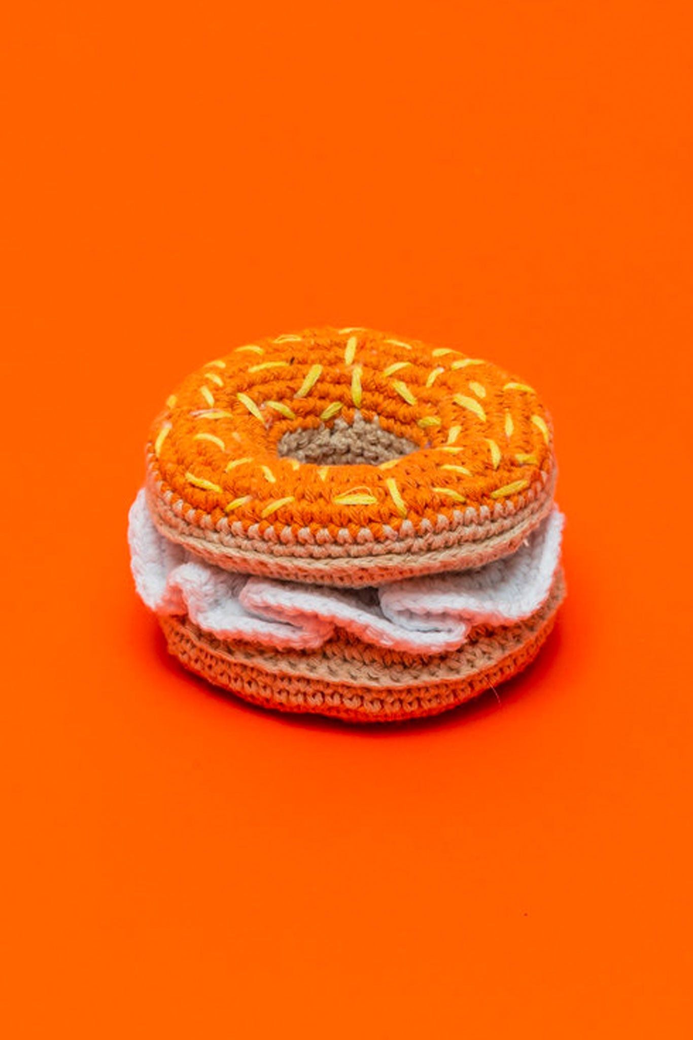 A crochet dog toy designed to resemble a bagel, with orange and white colors, on an orange background.