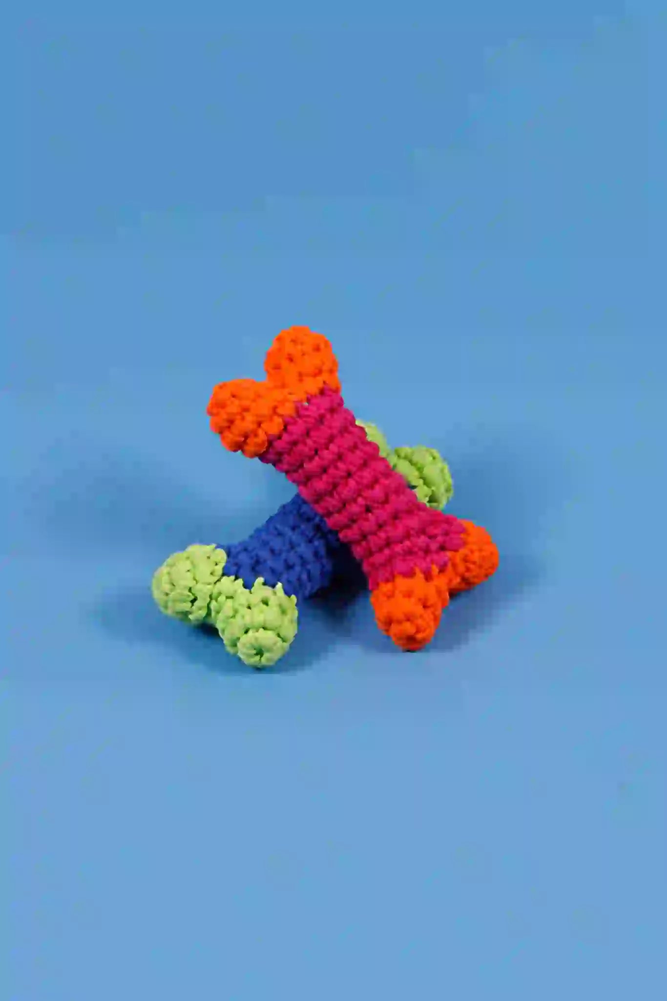A crochet dog bone toy in blue, green, and orange colors laid on a blue background.