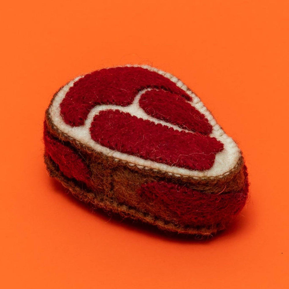 A felted wool dog toy shaped like a steak, with a red center and brown exterior, on an orange background.