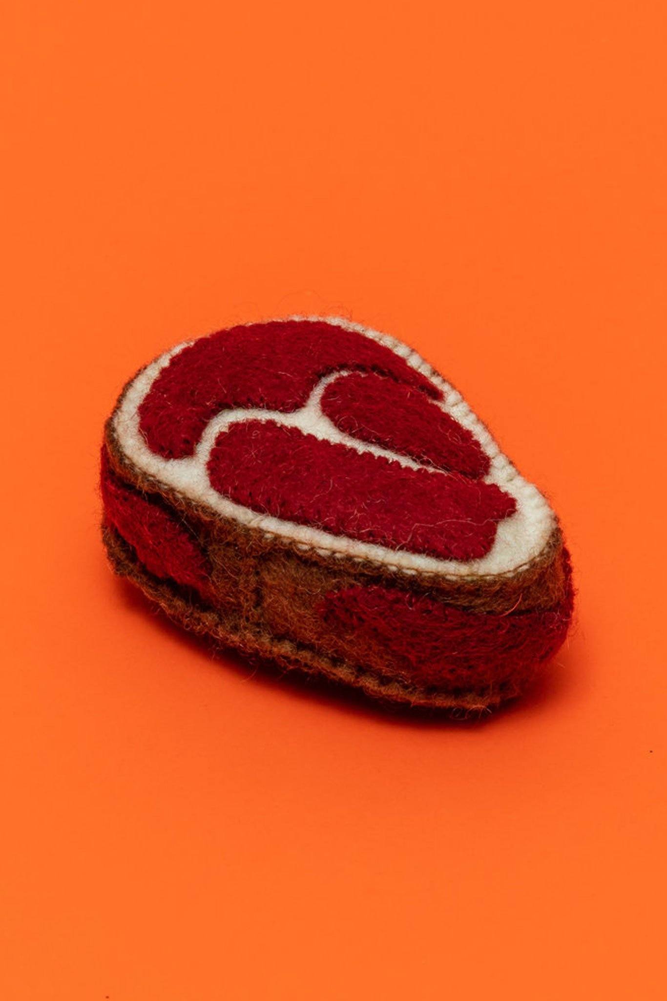 A felted wool dog toy shaped like a steak, with a red center and brown exterior, on an orange background.