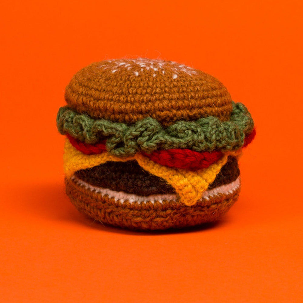 A hand knit dog toy resembling a hamburger with various colors for different components, placed against an orange background.