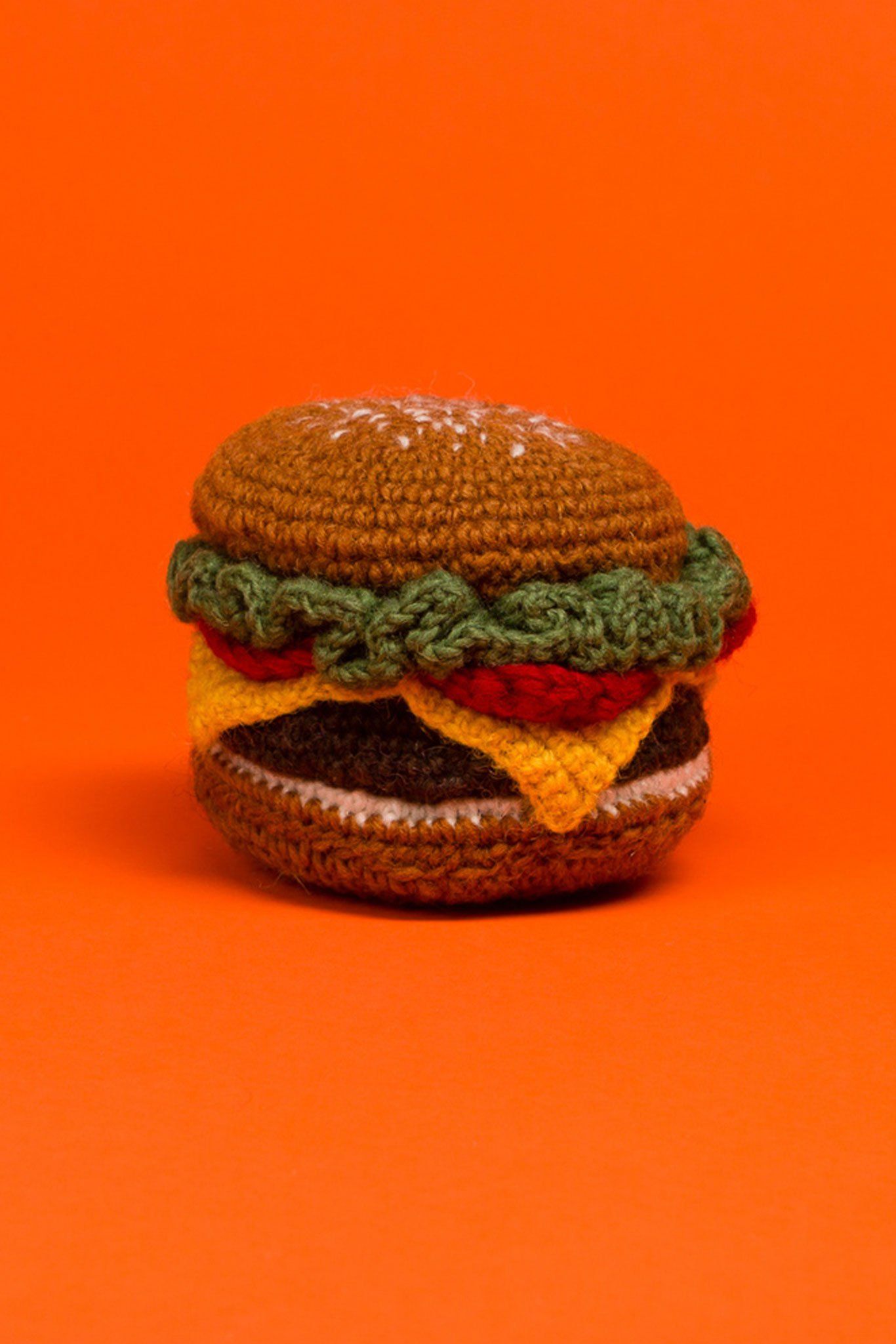 A hand knit dog toy resembling a hamburger with various colors for different components, placed against an orange background.