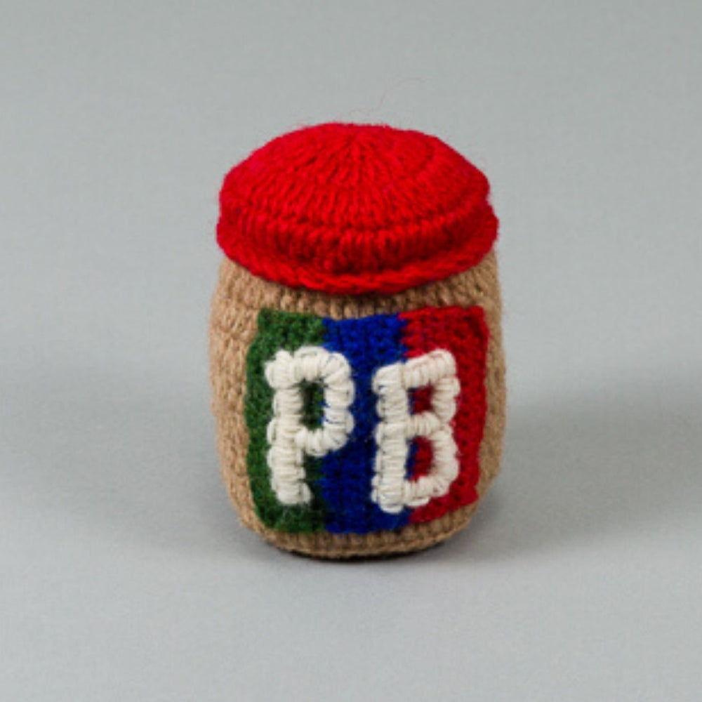 A hand knit, beige-colored dog toy shaped like a peanut butter jar with a red lid and the letters 'PB' on the side.