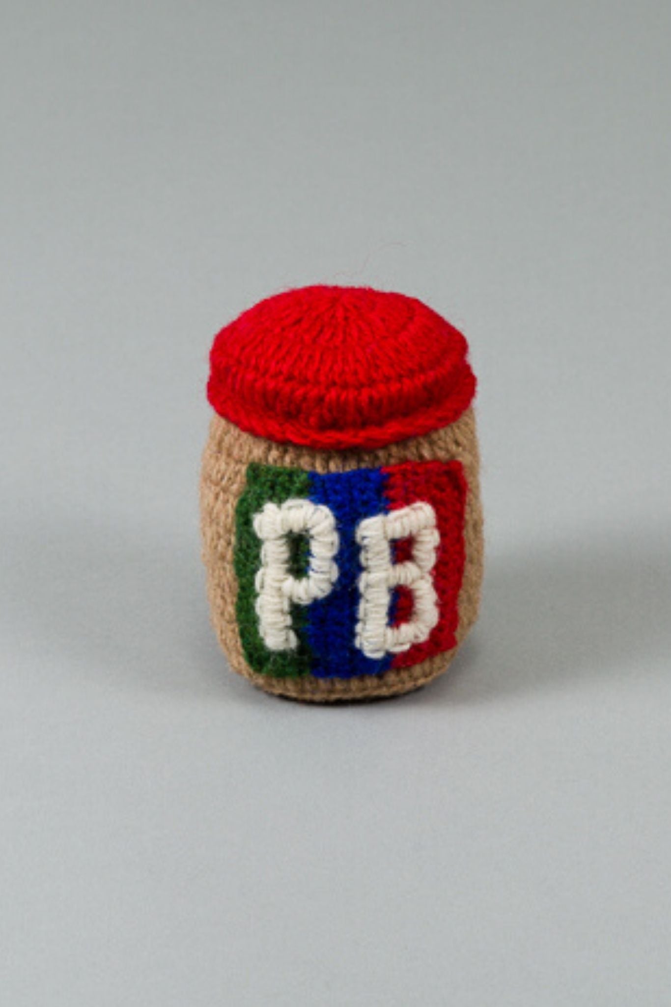 A hand knit, beige-colored dog toy shaped like a peanut butter jar with a red lid and the letters 'PB' on the side.
